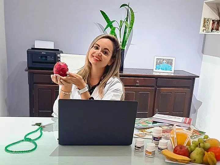 Natacha Ludkiewicz, de jaleco, segurando uma maçã à mesa do consultório, com notebook e frutas